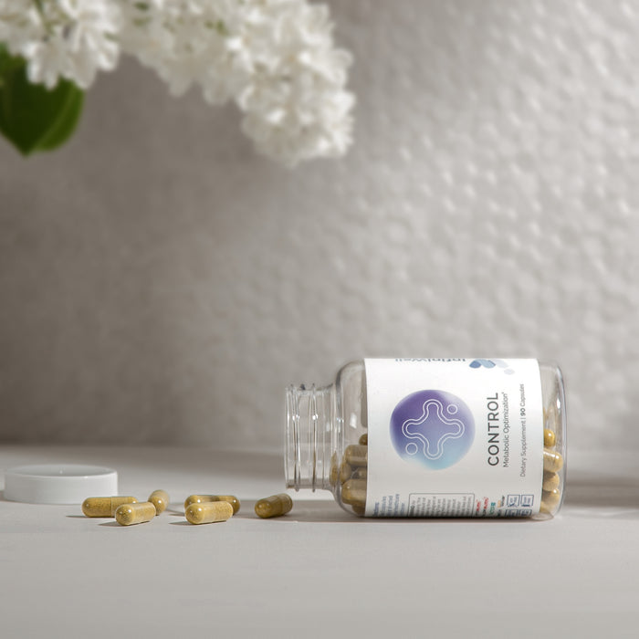 InfiniWell Control Metabolic Optimization supplement bottle with spilled capsules beside white flowers on a neutral surface.