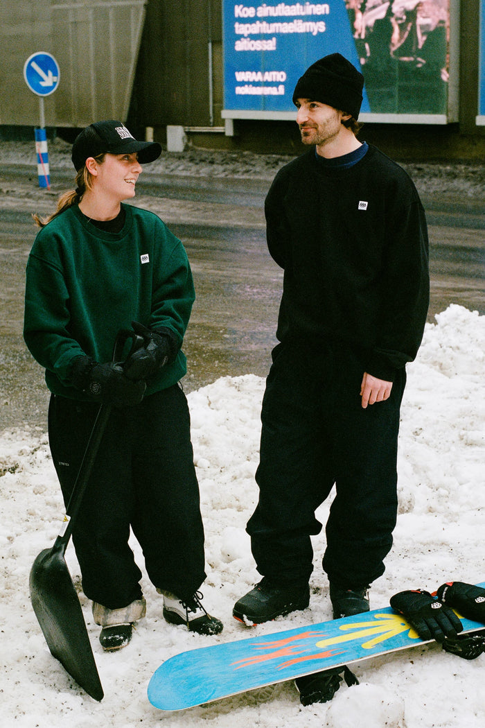Two people dressed in winter clothing, including the 686 Dojo® GORE-TEX Pant by 686, stand on snow; one holds a large black shovel while the other stands near a snowboard. Both appear to be talking and smiling.