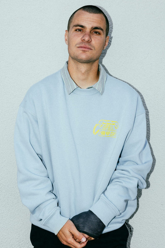 A young man with short dark hair stands against a light wall, wearing the 686 HUF Premium Heavyweight Crew Sweatshirt by 686 over a collared shirt, his hands clasped in front and his expression neutral.