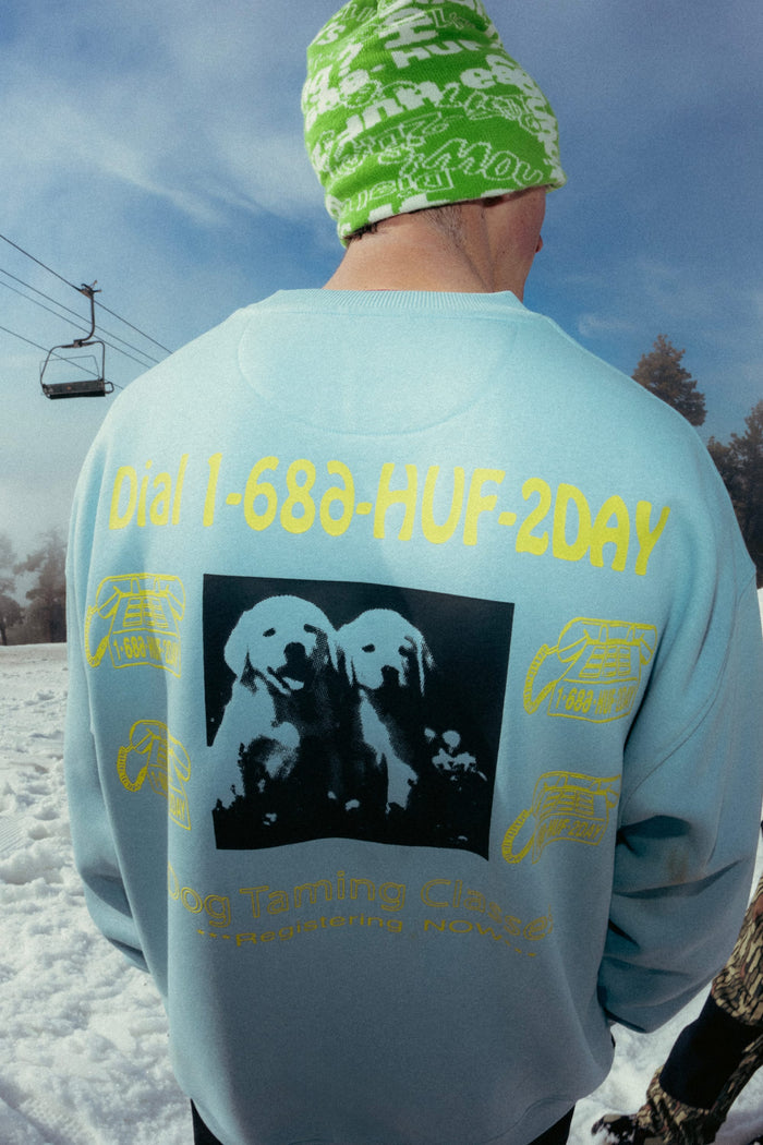 A person in a green beanie and blue 686 HUF Premium Heavyweight Crew Sweatshirt stands in the snow by a ski lift. The cotton polyester spandex blend sweatshirt features yellow dog graphics and ‚ÄúDial 1-888-HUF-2DAY‚Äù on the back.
