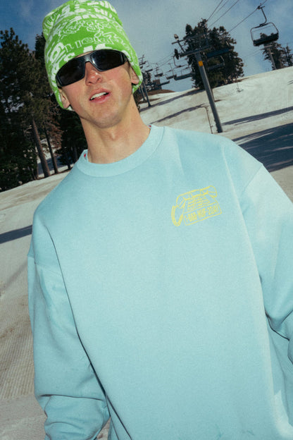 Wearing a 686 HUF Premium Heavyweight Crew Sweatshirt by 686, a green patterned beanie, and large sunglasses, a person stands outdoors on a snowy mountain near a ski lift, with trees and snow in the background.