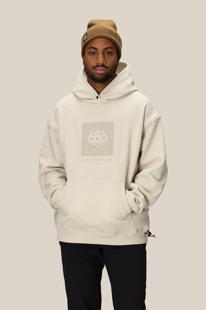 A man in a tan beanie and an oversized 686 Knockout Logo Premium Heavyweight Pullover Hoody stands against a plain beige background with his hands in the hoodie pocket.