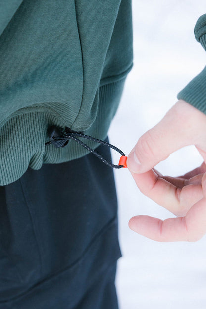 A person tightens the drawstring hem of a green 686 Knockout Logo Premium Heavyweight Pullover Hoody with their right hand. They‚Äôre wearing black pants, and the blurred white background suggests snow.