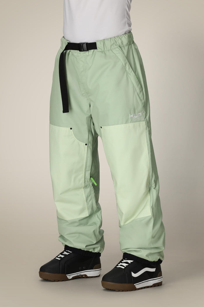 A person wears 686 Mens HUF Double H Shell Pant—loose, light green waterproof pants with a black belt and white HUF logo—paired with black and white skate shoes. The upper body is out of frame.