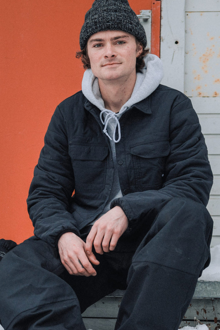 A young man in a 686 Men's Thermaldry™ Snap-Up Shirt sits outdoors before an orange and white wall, looking at the camera with a slight smile.