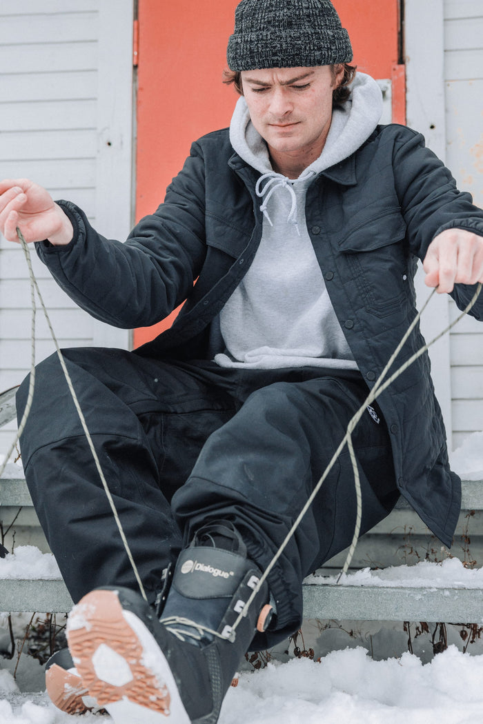 A person in winter gear sits on snowy steps, lacing black snow boots while wearing the 686 Men's Thermaldry™ Snap-Up Shirt. An orange door and white siding appear in the background.