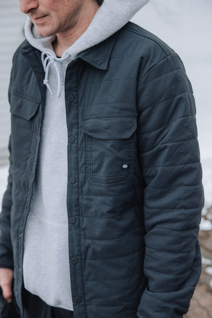 A person wearing the 686 Men's Thermaldry™ Snap-Up Shirt stands outdoors. Only the lower half of their face and upper body, layered beneath a grey hoodie, are visible in this cropped image.