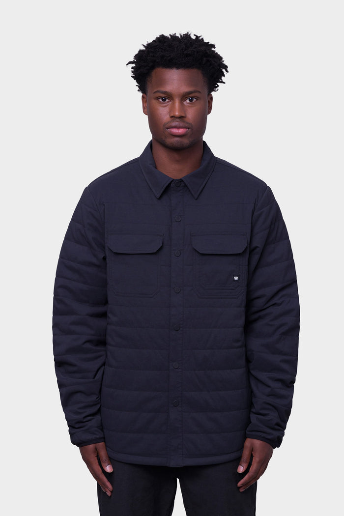 A person stands facing forward against a plain light gray background, wearing the 686 Men's Thermaldry™ Snap-Up Shirt by 686—a black quilted button-up with chest pockets.