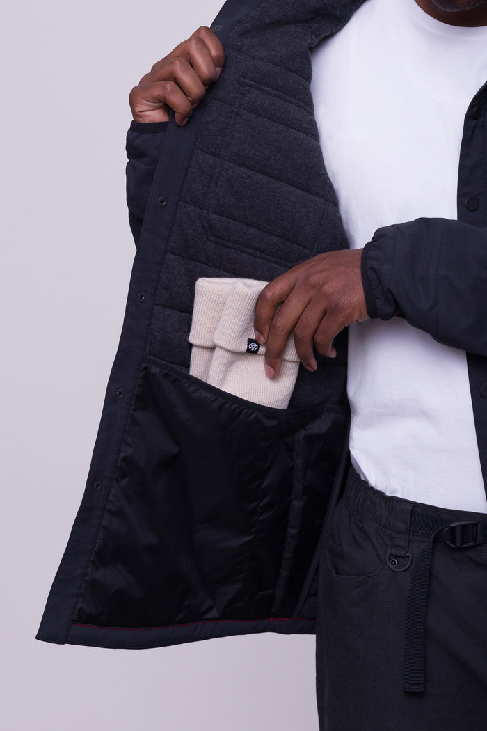 A person wearing the 686 Men's Thermaldry™ Snap-Up Shirt opens their black jacket, revealing a gray inner lining, and places a folded beige knit hat into an inner pocket.