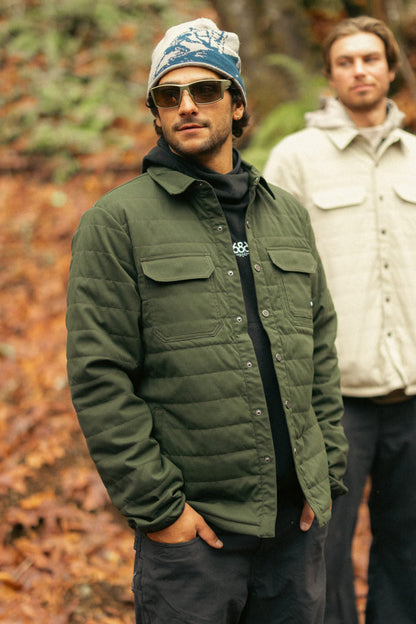 Two men are outdoors in autumn woods. The man in front wears sunglasses, dark pants, a printed beanie, and the 686 Men's Thermaldry™ Snap-Up Shirt by 686. The man behind wears a light jacket and stands slightly out of focus.