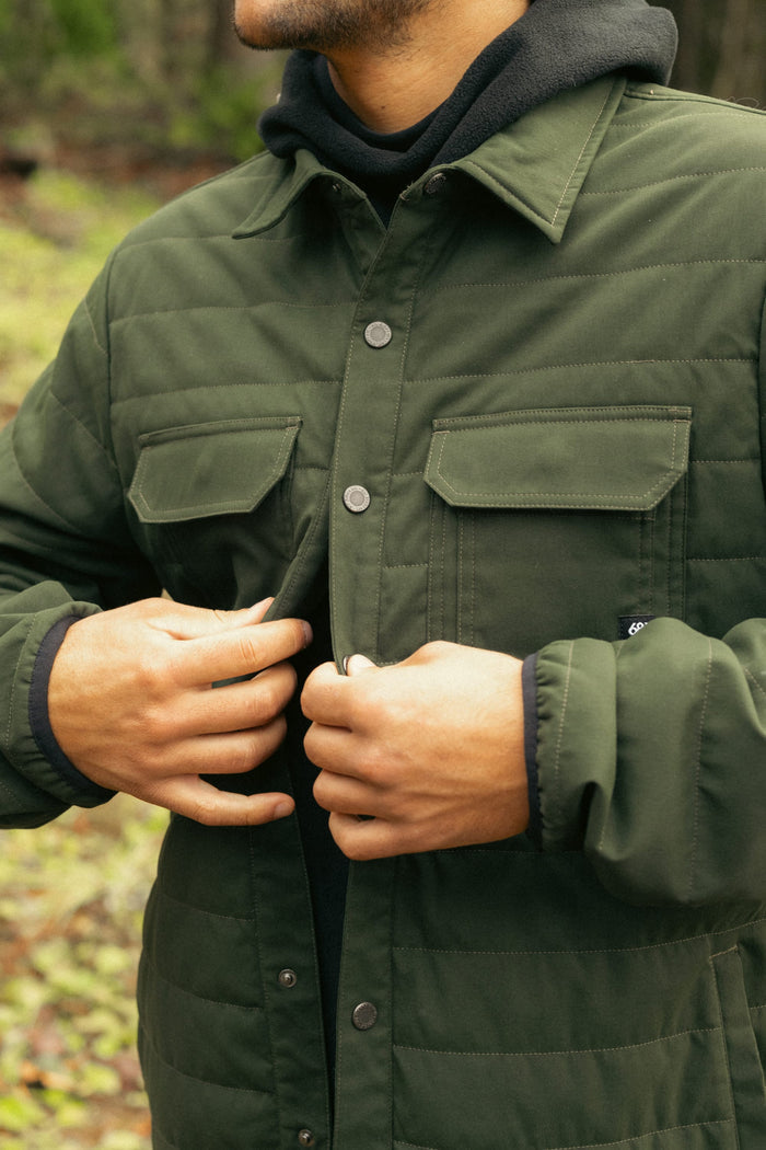 A person wearing the 686 Men's Thermaldry™ Snap-Up Shirt in dark green is buttoning it up over a black hoodie outdoors. The photo highlights the torso and hands, set against a softly blurred natural background.