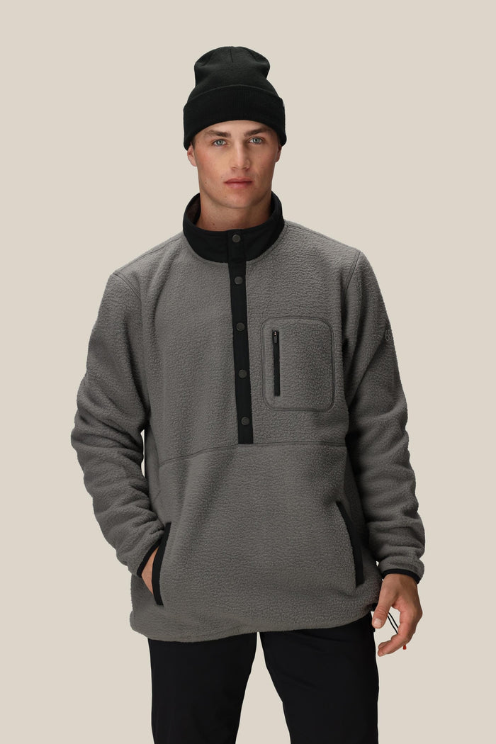A young man wearing a 686 Mens Tioga Fleece Pullover‚Äîfeaturing a button collar and chest pocket‚Äîstands with one hand in his pocket against a plain beige background.