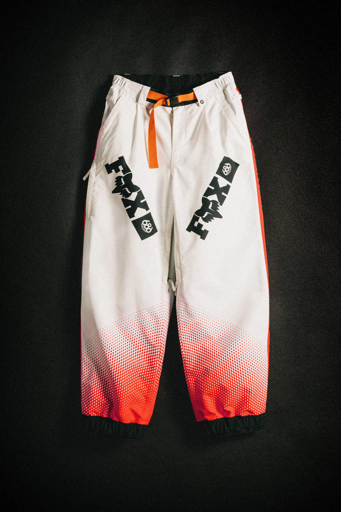 The 686 Women's Fox Racing Shell Pant by 686 features baggy white legs with bold black FMAX logos, orange side panels and belt, plus a red gradient near the ankle cuffs, shown against a dark background.