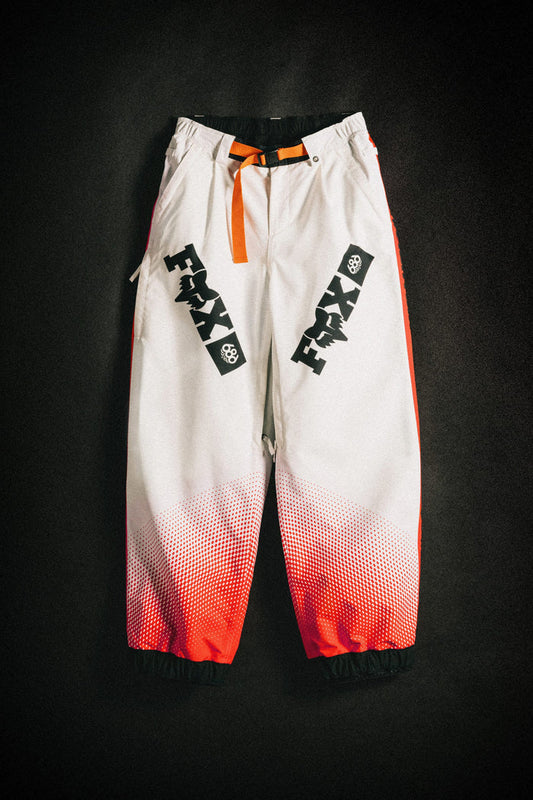 The 686 Women's Fox Racing Shell Pant by 686 features baggy white legs with bold black FMAX logos, orange side panels and belt, plus a red gradient near the ankle cuffs, shown against a dark background.