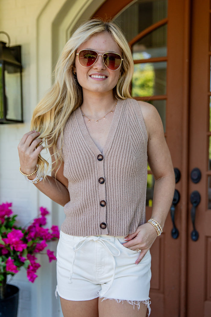 A woman wearing a sleeveless ribbed knit button-front vest in a warm taupe-beige color, paired with white shorts and sunglasses, standing in front of a brown wooden door.