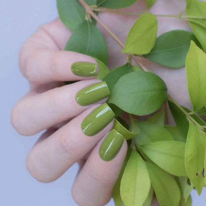 Hand with green nail polish holding a leafy branch against a light purple background.