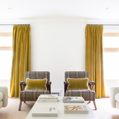 Golden chartreuse velvet curtains on metal rods in front of illuminated windows with blue and yellow striped chairs with Golden chartreuse velvet pillows on them and a white coffee table with books spread on it