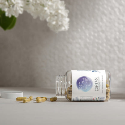 InfiniWell Control Metabolic Optimization supplement bottle with spilled capsules beside white flowers on a neutral surface.