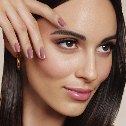 A person with long dark hair touches their face, highlighting LONDONTOWN's Crowning Crumpet (Professional) sheer nude nail polish. Their subtle makeup pairs perfectly with their gold hoop earrings, creating an effortlessly chic look.