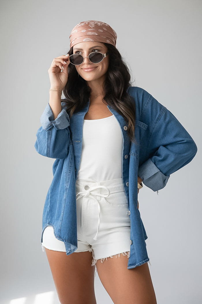 A woman posing in a styled outfit featuring a blue denim button-up shirt, white tank top, white shorts, a pink patterned headband, and sunglasses against a light gray studio background.