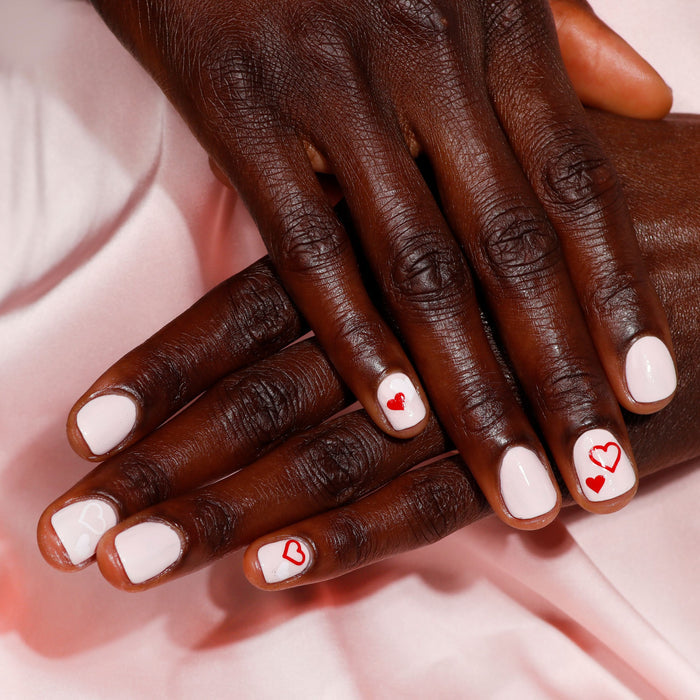 Hands with LONDONTOWN's Invisible Crown white nail polish and red heart designs rest on silky, sheer pink fabric.