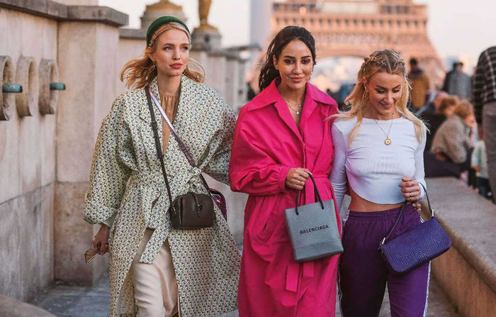 Three women walking together on a street, with one in a pink coat and another in a purple outfit, in an urban setting.