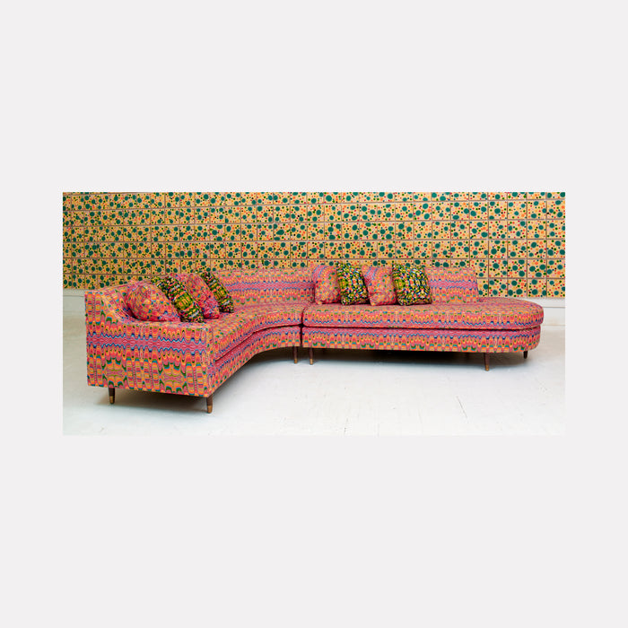 The artwork 'Piece of Cake 912' by Lori Kirkbride features a vibrant sectional sofa in coral pink with intricate multicolored geometric patterns. The L-shaped couch is set against a retro-inspired wallpaper with green and gold polka dots. The acrylic and resin artwork captures mid-century modern furniture design with decorative throw pillows matching the upholstery pattern.