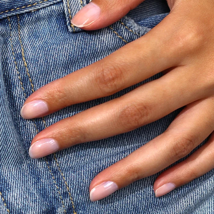A hand with nails polished in LONDONTOWN’s Pink Illuminating Nail Concealer, featuring a milky pink finish, rests on blue denim jeans.