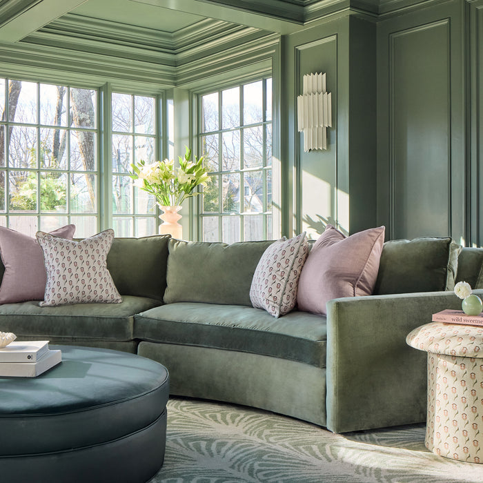 blue green couch with pink pillows and abstract floral pink pillows on the couch, a peacock bleu ottoman in front of the couch with books on it, abstract pink floral fabric side table with a book on it next to the couch in a living room painted light green with illuminated windows behind the couch