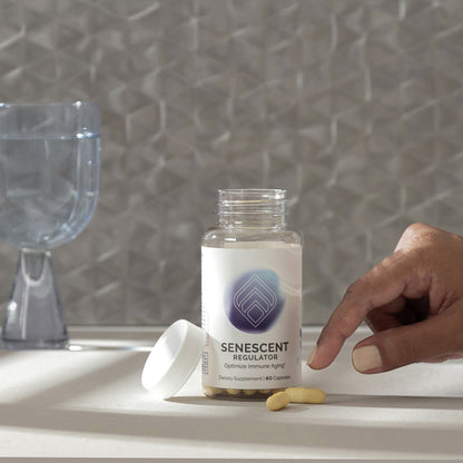 InfiniWell Senescent Regulator dietary supplement bottle with open lid, capsules, hand, and glass of water on a neutral surface.