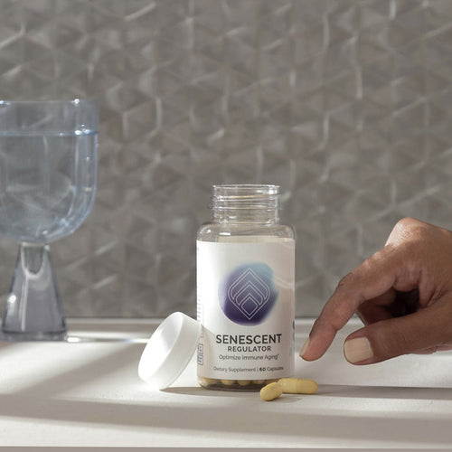 InfiniWell Senescent Regulator dietary supplement bottle with open lid, capsules, hand, and glass of water on a neutral surface.