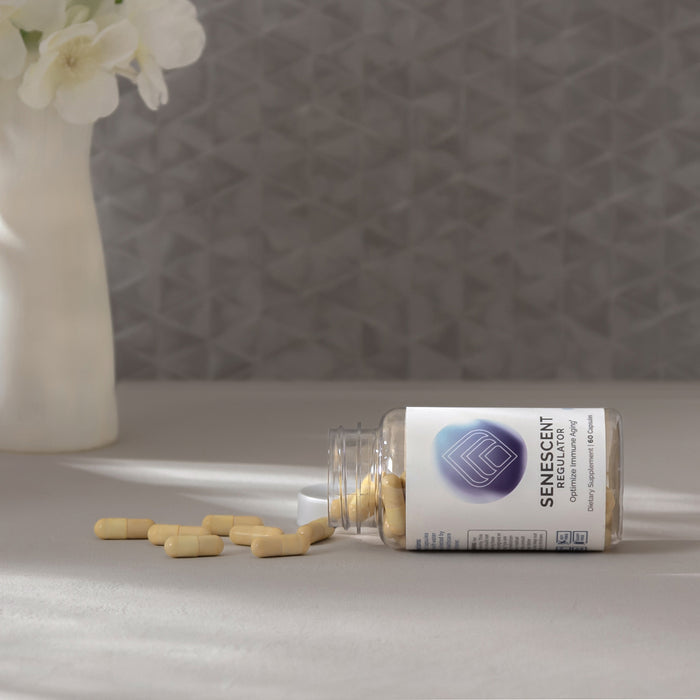 InfiniWell Senescent Regulator dietary supplement bottle with spilled capsules on a light surface beside a white flower vase.