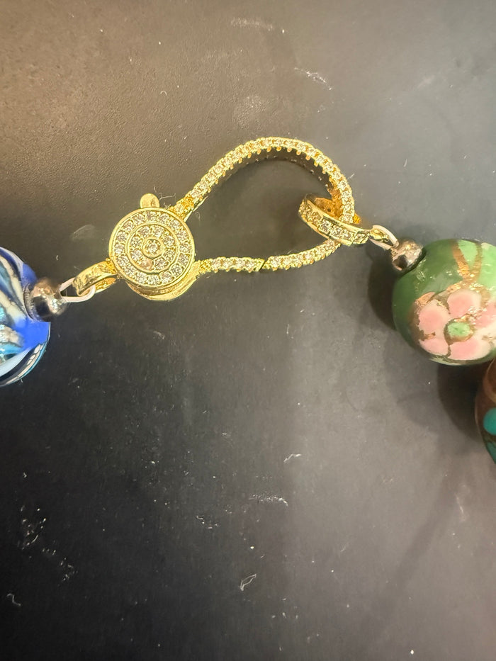 A close-up of a gold-toned beaded necklace or bracelet with a central round gold medallion and textured gold chain, flanked by colorful beads including blue, green, and pink floral designs, on a dark surface.