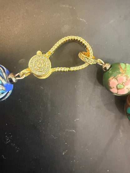 A close-up of a gold-toned beaded necklace or bracelet with a central round gold medallion and textured gold chain, flanked by colorful beads including blue, green, and pink floral designs, on a dark surface.