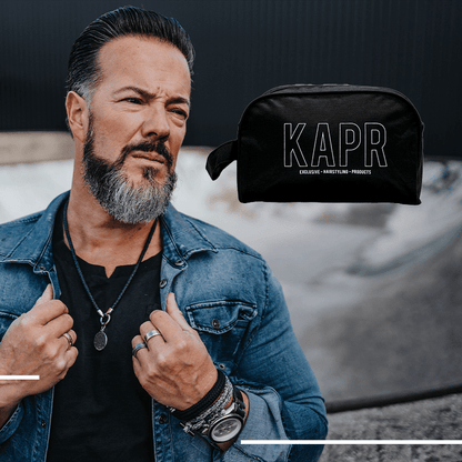 Man met baard poseert naast zwarte KAPR toilettas – premium accessoire voor moderne barbers.